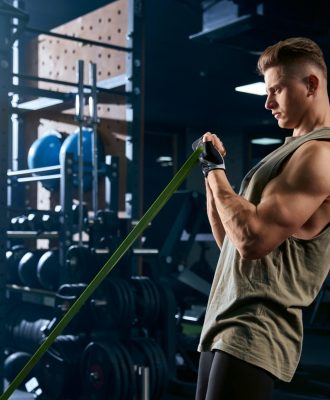 Portrait of muscular bodybuilder training arms with fitness resistance bands. Side view of man with perfect body training biceps in gym in dark atmosphere. Concept of bodybuilding.
